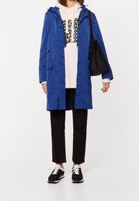 Lång blå regnjacka med huva, två framfickor, bärs över en ljus Sweatshirt, ihop med svarta byxor och svarta sneakers med vita snörningar.