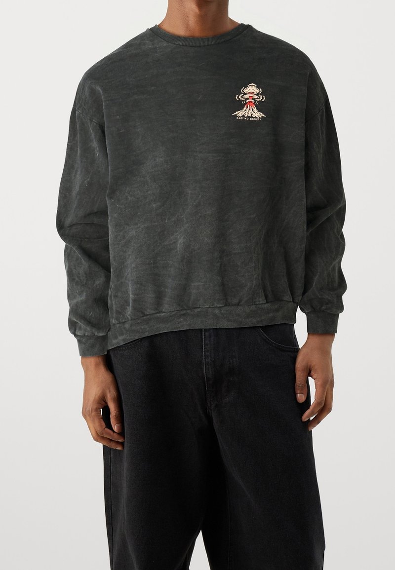 Personne portant un sweat-shirt noir à manches longues lavé avec un petit motif d’éruption volcanique sur le côté gauche de la poitrine et un pantalon noir ample.