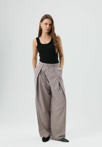 Zwart geribd tanktopje, lichtgrijze wijde broek met hoge taille en plooien aan de voorkant, model poseert met handen in de zakken, zwarte platte schoenen.