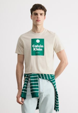 Fiatal férfi bézs Calvin Klein pólóban zöld logóval, világoskék farmernadrágban és zöld csíkos pulóverrel, amelyet a derekán kötött meg.