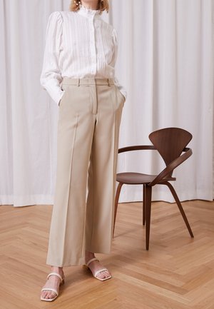 Kvinde i hvid langærmet bluse og beige bukser med høj talje og vide ben står indendørs nær en moderne træstol.