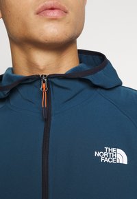 Niebieska kurtka z kapturem na zamek błyskawiczny, z pomarańczowymi akcentami i białym logo na klatce piersiowej. Gładka tekstura, sportowy design.