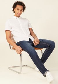 Camicetta bianca a maniche corte in lino, jeans blu in denim, seduto su una sedia in cromato con seduta in legno, sneakers bianche, tatuaggio sottile sull'avambraccio.