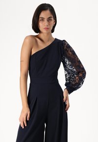 Navy jumpsuit featuring an asymmetrical one-shoulder design, long lace balloon sleeve, and pleated front for added texture.