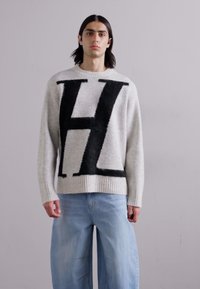 Szürke kötött pulóver, nagy fekete 'H' logóval az elején, bordás mandzsettákkal és aljjal. Világoskék, bő szárú farmernadrággal viselve.