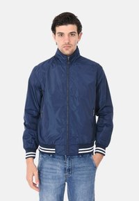Giacca bomber leggera blu navy con colletto alto, chiusura con zip, tasche laterali e polsini e orlo a righe. Tessuto liscio e lucido.