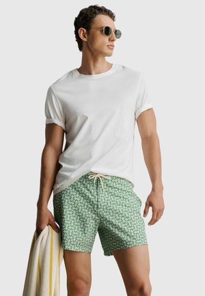 Ein Mann mit Sonnenbrille, weißem T-Shirt und grünen gemusterten Badeshorts hält ein gestreiftes Handtuch und steht vor einem schlichten Hintergrund.
