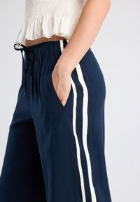 Navy blue wide-leg pants with a drawstring waist, featuring cream vertical stripes on both sides and side pockets. Smooth fabric texture.