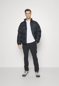 Doudoune noire avec un col montant et fermeture éclair, portée sur un t-shirt blanc. Associée à un pantalon cargo noir et des baskets multicolores.