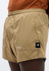 Shorts de sport marron clair en tissu lisse, dotés d'une ceinture élastique, de poches et d'accents gris ; marqués d'un patch logo noir.