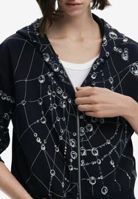 Sudadera con cremallera de color navy que presenta un patrón de círculos y líneas interconectadas, con una textura suave y acentos blancos, llevada sobre una camiseta blanca.