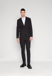 Black suit with slim-fit design, featuring a single-button jacket, notch lapels, and flat-front trousers. White shirt with black buttons. Black shoes.