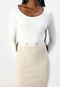 Haut à manches longues blanc avec un décolleté arrondi, associé à une jupe beige texturée dotée de deux boutons décoratifs à la taille.