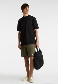 T-shirt en coton noir avec un petit logo, short vert olive et chaussures slip-on de couleur claire, tenant un sac fourre-tout noir. Design simple et décontracté.
