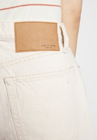 Jeans de mezclilla blanca con un parche de cuero marrón que presenta el texto "rag & bone NEW YORK." Costuras visibles y detalle del bolsillo trasero.