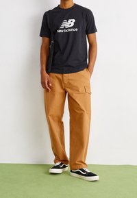 Czarny bawełniany T-shirt z białym logo "New Balance", w połączeniu z beżowymi spodniami cargo i czarno-białymi sneakersami, stojący na zielonej podłodze.