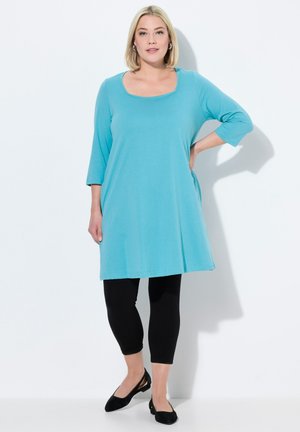 En tekanfarvet tunika kjole med rund halsudskæring og trekvartærmer, parret med sorte leggings og sorte slingback flade sko.