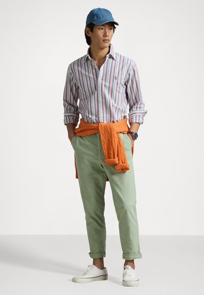 Randig skjorta, gröna byxor, orange tröja knuten runt midjan, denimkeps och vita sneakers. Släta texturer och avslappnad design.