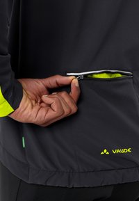 Una mano che apre la zip di una tasca nera di una giacca Vaude, con l'interno giallo neon e il logo visibile sul lato inferiore destro.