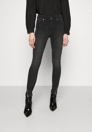 Kobieta ubrana w czarne, obcisłe jeansy z wysokim stanem i szwami na kolanach, czarne botki na zamek przy kostce oraz czarną bluzkę z długim rękawem z guzikami.