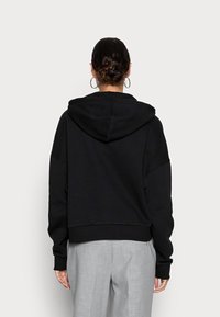 Black cropped hoodie with a drawstring hood, oversized sleeves, and ribbed hem; worn with light gray trousers. Soft fabric texture.
