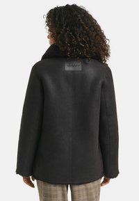 Zwarte schapenvacht jas met een grote kraag en GANT logo patch. Glad materiaal met een precies passende snit; zichtbare naden; gedragen over een geruite broek.