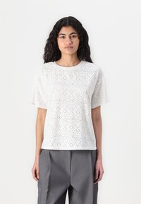 Blouse blanche à manches courtes en tissu léger avec des détails en œillets formant un motif, encolure ronde et coupe décontractée.