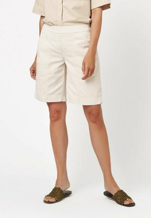 Beige Baumwollshorts mit einem breiten elastischen Bund, ausgestattet mit Taschen und einem geraden Schnitt. Kombiniert mit gewobenen dunkelgrünen Sandalen.