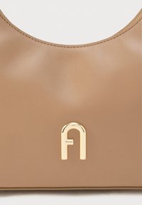 Brązowa skórzana torba o gładkiej fakturze, z zaokrąglonym górnym brzegiem i złotym metalowym elementem z stylizowanym logo w kształcie litery na przodzie.