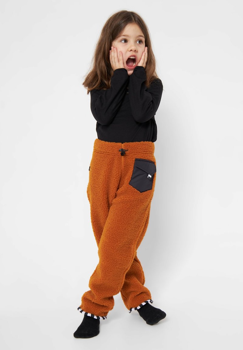 Brune fleecebukser med en sort lomme, elastisk talje og rynkede manchetter. Barnet har en sort langærmet bluse på, hænderne på ansigtet.