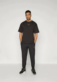 Camiseta negra de manga corta con logo en el pecho, combinada con pantalones joggers negros que tienen el logo en la pierna izquierda y zapatillas deportivas negras.