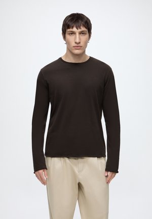 Joven con una camisa negra de manga larga y pantalones beige, de pie frente a un fondo claro y sencillo, mirando hacia adelante.