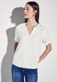 Blusa bianca a maniche corte con scollatura a V, realizzata in tessuto morbido, caratterizzata da maniche arrotolate e una leggera lucentezza, abbinata a jeans blu.