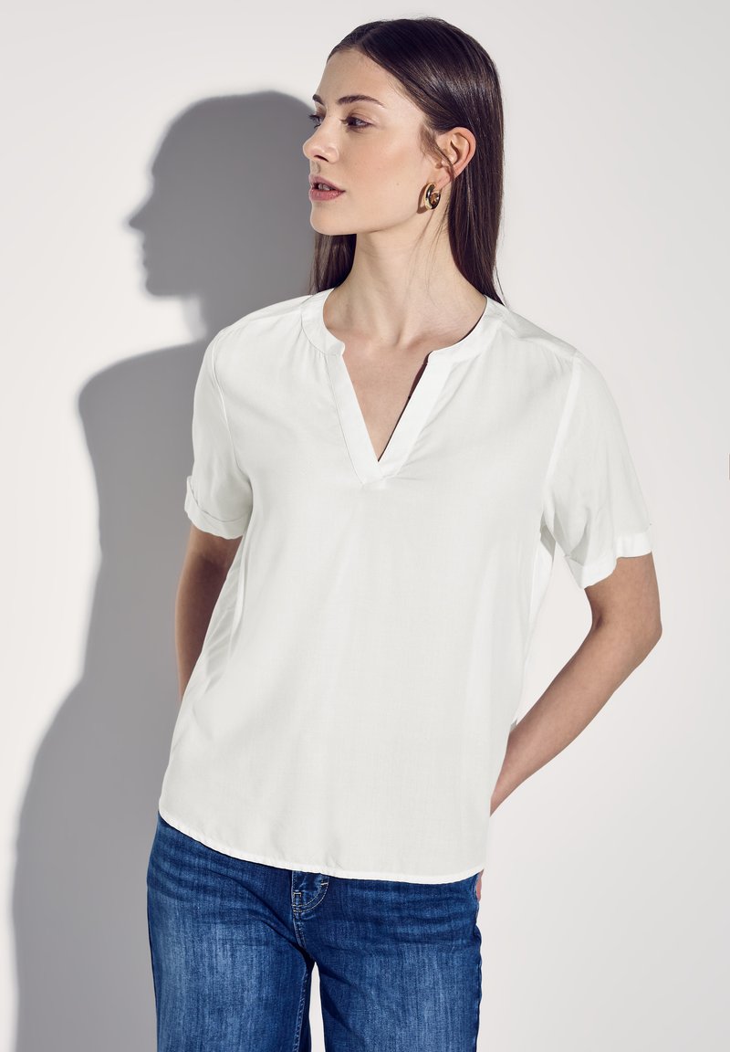 Blusa bianca a maniche corte con scollatura a V, realizzata in tessuto morbido, caratterizzata da maniche arrotolate e una leggera lucentezza, abbinata a jeans blu.