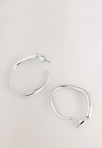 Silver hoop earrings with a fluid, wavy design, featuring a cylindrical post and a smooth, reflective surface.