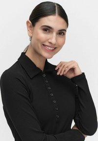 Musta ribbattu pitkähihaisuus polo-paita, jossa on kauluspaita ja nappilista. Sulavaliukuinen pinta, jossa on hienovaraiset pystysuuntaiset viivat.