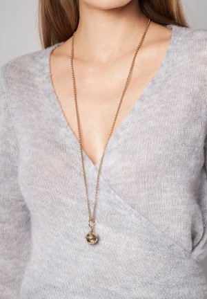 Grå, blød striktrøje med en dyb V-udskæring og et guld kædetrøje med en rund vedhæng. Vedhænget har intrikate udskårne mønstre.