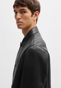 Black leather jacket featuring a button closure, notched collar, and smooth texture, shown on a male model from a side profile.