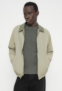 Νεαρός άνδρας με κοντά σγουρά μαλλιά, φοράει ανοιχτό μπεζ μπουφάν με φερμουάρ πάνω από γκρι πλεκτό πουλόβερ και μαύρο παντελόνι, με τα χέρια στις τσέπες, κοιτάζοντας προς τα κάτω.