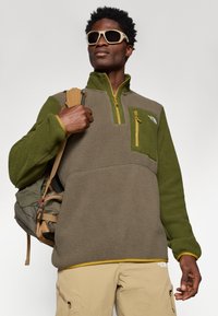 Pullover in pile verde e grigio con mezzzip, tasca frontale e dettagli gialli. Il modello indossa uno zaino e occhiali da sole.