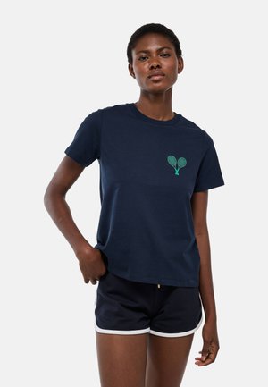Femme portant un t-shirt bleu marine avec un logo de raquettes de tennis vertes et un short bleu marine assorti avec un bord blanc, debout la main dans la poche.