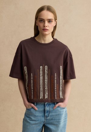 Joven con camiseta marrón holgada con bordado vertical de plantas en crema y marrón, combinada con vaqueros azules, manos en los bolsillos.