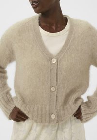 Beige Strickjacke mit V-Ausschnitt, fünf Knöpfen, gerippten Bündchen und Saum. Hergestellt aus weichem, flauschigem Material. Geprägte Struktur erkennbar.