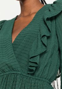 Abito avvolgente verde con tessuto texturizzato caratterizzato da strisce verticali. Le spalle arricciate e il punto vita stretto arricchiscono il suo design.