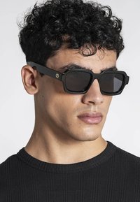 Hombre con cabello rizado negro, lleva gafas de sol cuadradas negras y una camiseta negra acanalada con cuello redondo, mirando hacia la derecha.