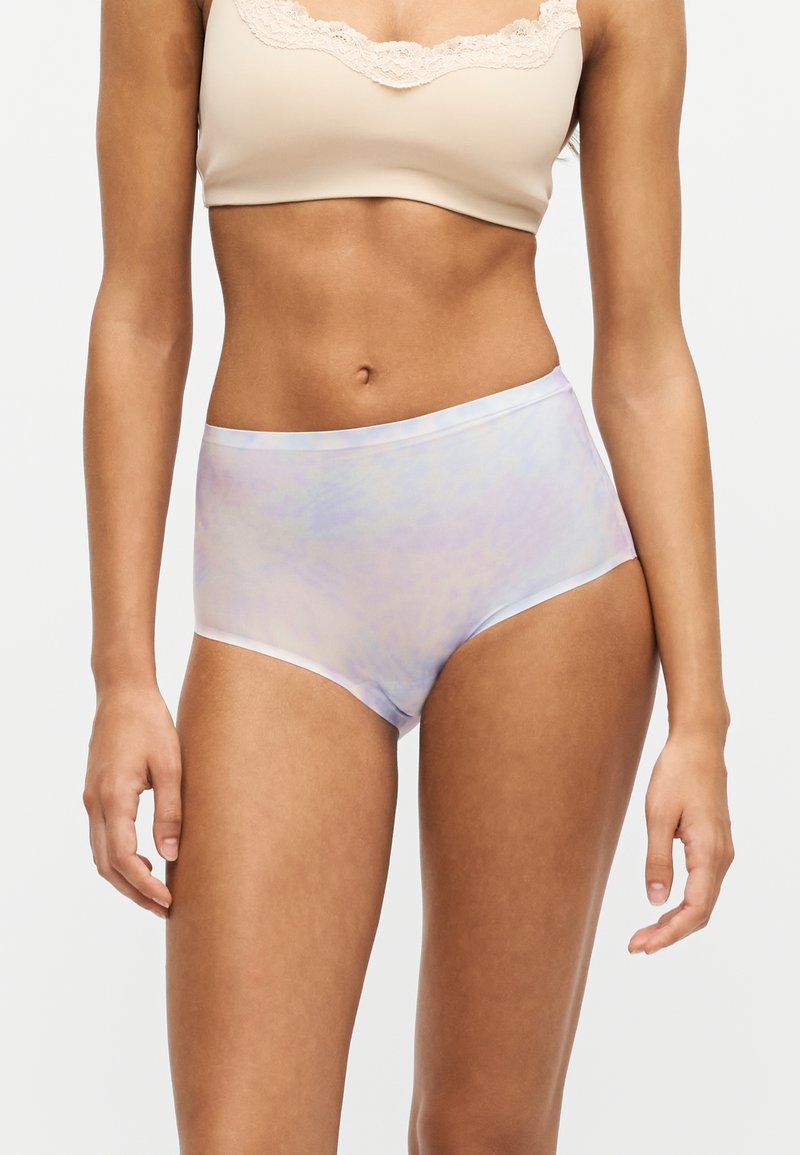 Femme portant des sous-vêtements tie-dye pastel taille haute et un soutien-gorge beige à bordure en dentelle, debout devant un fond blanc uni.