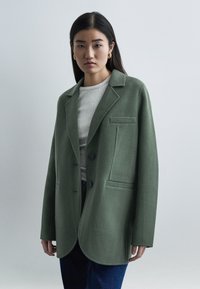 Νεαρή γυναίκα με μακριά σκουρόχρωμα μαλλιά φοράει υπερμεγέθη πράσινο μάλλινο σακάκι, κρεμ ριμπ πλεκτό τοπ, μπλε τζιν και χρυσούς κρίκους σκουλαρίκια σε απλό φόντο.