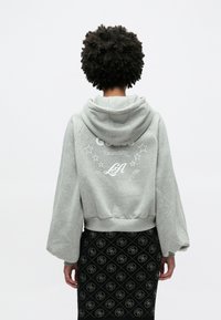 Γκρι κοντοκομμένο φούτερ με κουκούλα και μακριά, φαρδιά μανίκια. Διαθέτει λευκή κεντητή επιγραφή «GUESS» και αστέρια στην πλάτη. Συνδυασμένο με μαύρη φούστα με σχέδιο.