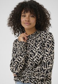 Blouse à manches longues avec encolure froncée, présentant un motif abstrait noir et crème, fabriquée en tissu léger avec une légère texture.