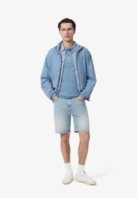 Chaqueta azul claro sobre un polo azul, combinada con pantalones cortos de mezclilla claros. Presenta detalles del logo, zapatillas blancas y calcetines blancos.